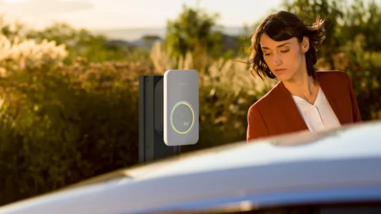 Eine Frau steht im Freien neben einer an der Wand montierten Fronius Wattpilot Ladestation vor einem sonnigen, herbstlichen Hintergrund.