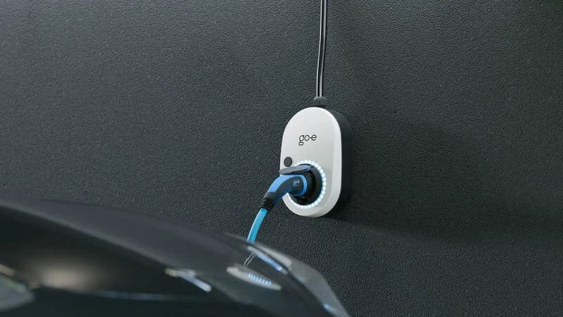 Weißer go-e Charger an einer dunklen Wand montiert. Die smarte Wallbox lädt über ein blaues Ladekabel ein Elektroauto, der LED-Ring der Ladestation leuchtet.